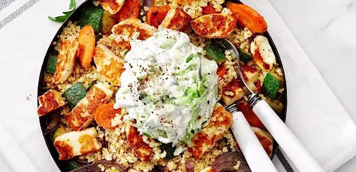 Halloumisallad med stekta halloumibitar, rostad zucchini, morot och rödlök på en bädd av gryn, toppad med en klick tzatziki, serverad i en skål med två bestick