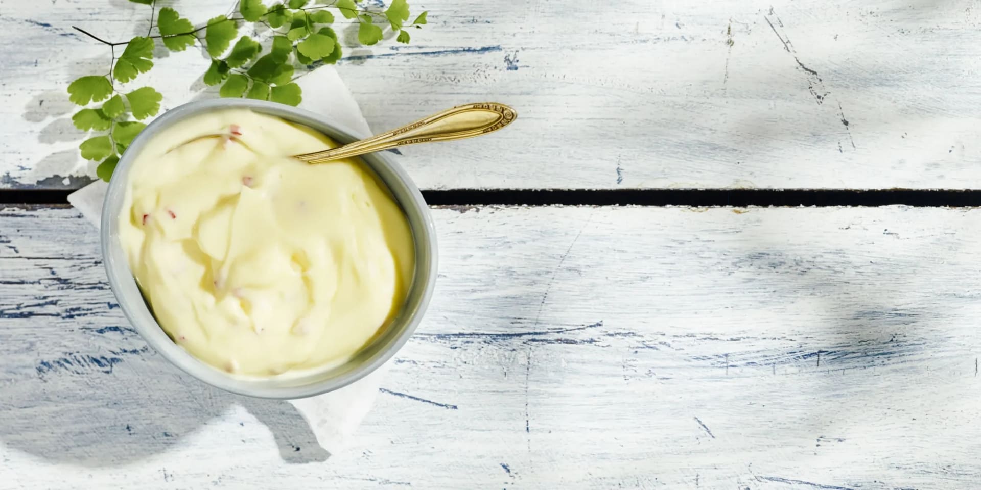 Aioli i en skål med en gulddsked i, skålen står på ett vitlaserat träbord. Ovanför skålen ligger en knippe gröna blad. 