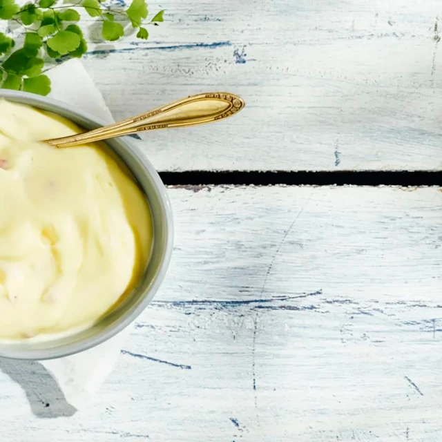 Aioli i en skål med en gulddsked i, skålen står på ett vitlaserat träbord. Ovanför skålen ligger en knippe gröna blad.