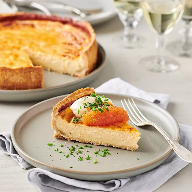 En bit cheesecake med gräddfyllning, toppad med gräslök och laxrom, serverad på en tallrik. I bakgrunden syns en hel tårta.