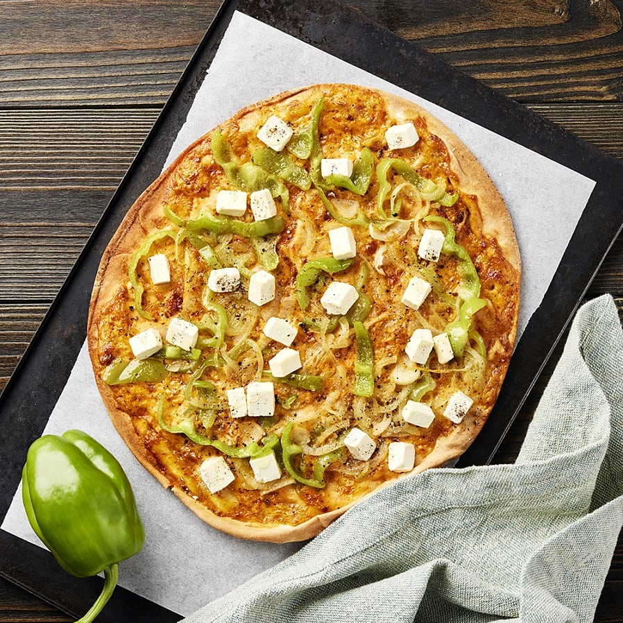 Pizza med grön paprika, lökar och fetaost på en bakplåt, med en grön paprika bredvid och en duk i hörnet.