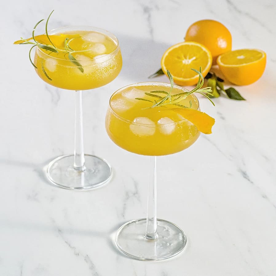 Två cocktailglas med is, apelsindryck och rosmarinkvistar, samt apelsiner halverade i bakgrunden.