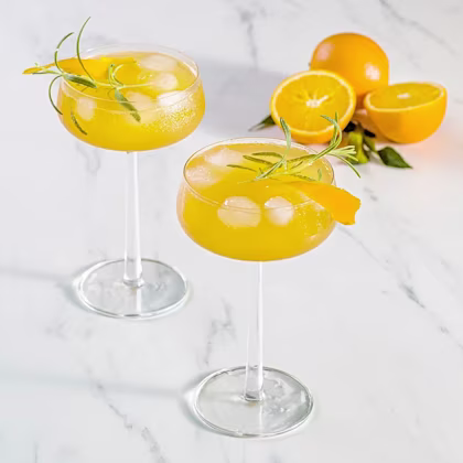 Två cocktailglas med is, apelsindryck och rosmarinkvistar, samt apelsiner halverade i bakgrunden.