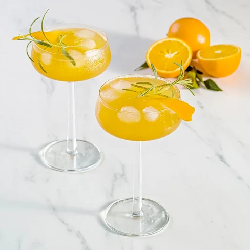Två cocktailglas med is, apelsindryck och rosmarinkvistar, samt apelsiner halverade i bakgrunden.