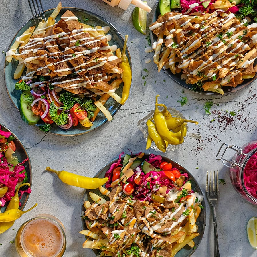 Tre tallrikar med gyros, pommes frites, färska grönsaker och sås, omgivna av jalapeños och en burk med inlagd rödkål.