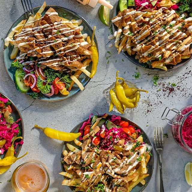 Tre tallrikar med gyros, pommes frites, färska grönsaker och sås, omgivna av jalapeños och en burk med inlagd rödkål.