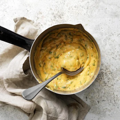 En liten kastrull med Chilibearnaisesås, kastrullen står på ettbord av grå sten, en beige linneservett ligger till vänster om kastrullen. En sked är i såsen.