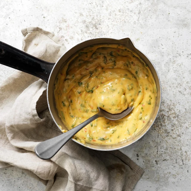 En liten kastrull med Chilibearnaisesås, kastrullen står på ettbord av grå sten, en beige linneservett ligger till vänster om kastrullen. En sked är i såsen.