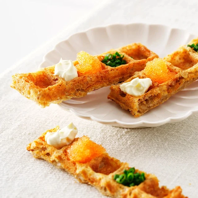 Två bitar våfflor på en assiett och en bit på sidan om på en linne duk. Toppade med små klickar av smetana, löjrom och gräslök.