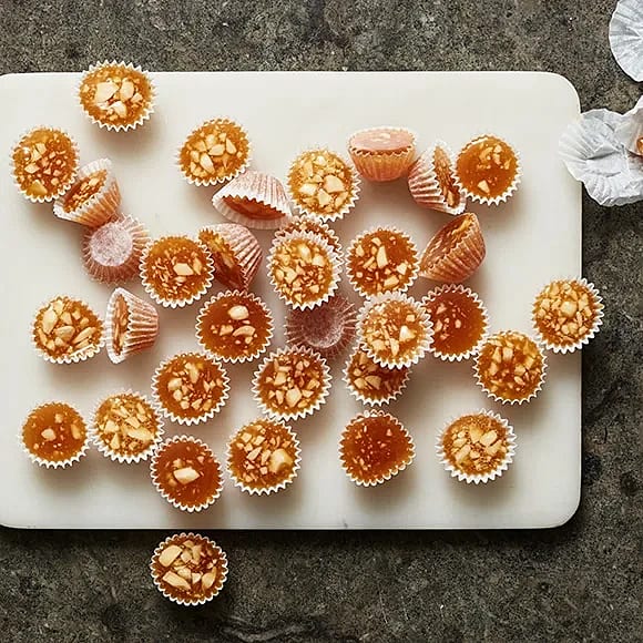 Små godisbitar i pappersformar, spridda på en skärbräda, med en glänsande yta och nötter synliga i några av dem.