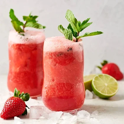 Två röda jordgubbsdrinkar med mynta som garnering, omgivna av is och limehalvor.