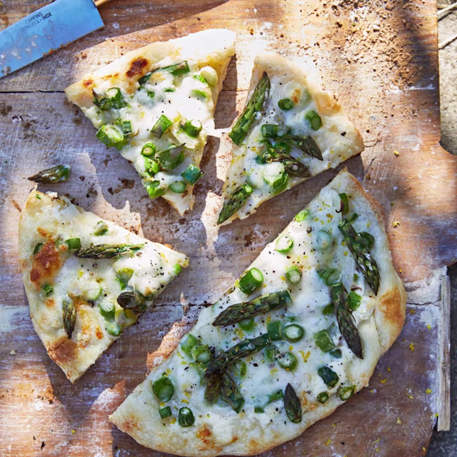 Pizza med vit ost och grön sparris i bitar, uppskuren i fyra trekanter på en träbräda.