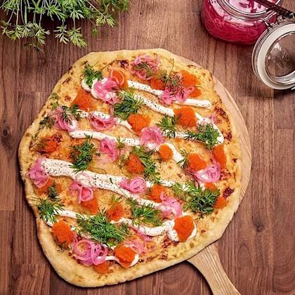 Tunnbrödspizza med crème fraiche, löjrom, dill och picklad rödlök på träbricka med burk och skål bredvid.