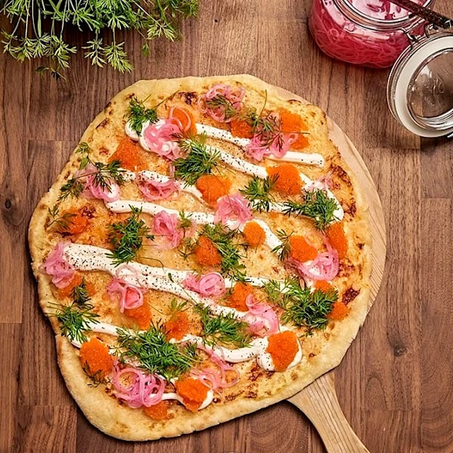 Tunnbrödspizza med crème fraiche, löjrom, dill och picklad rödlök på träbricka med burk och skål bredvid.