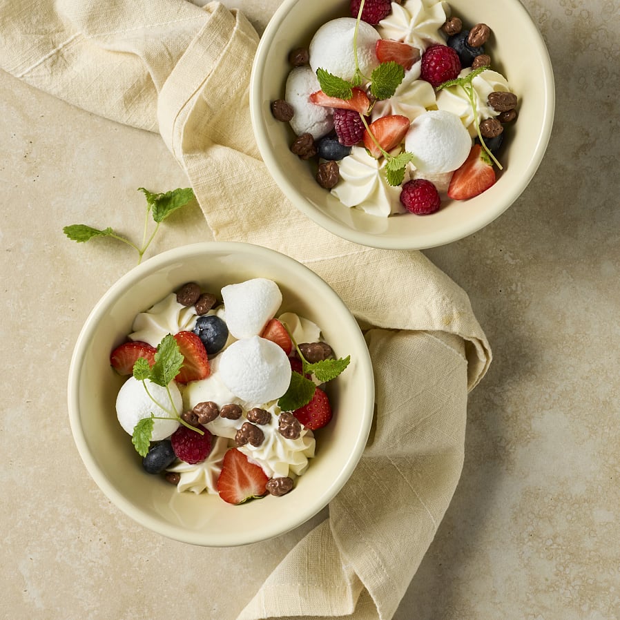 Två skålar med grädde, glass och maräng. Toppade med bär, chokladpuffar och gröna blad. 