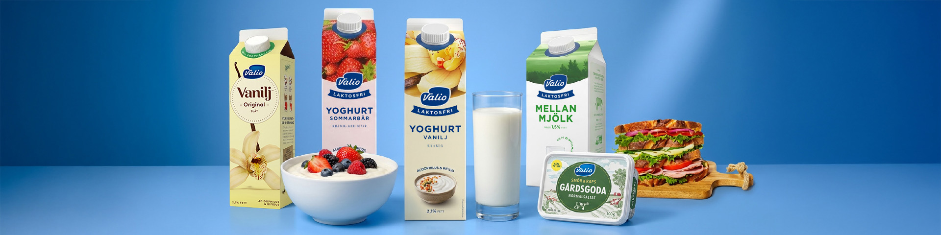 Förpackningar med Valio laktosfri vaniljdryck, yoghurt sommarbär och vanilj samt mellanmjölk, tillsammans med skål med yoghurt och bär, glas mjölk, smörgåsar och ask med smör- och rapsolja. Klicka för att handla.