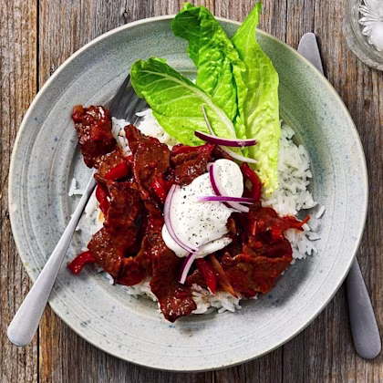 Köttgryta med paprika och lök serverad på ris, garnerad med sallad och en klick gräddfil, på ett träbord.