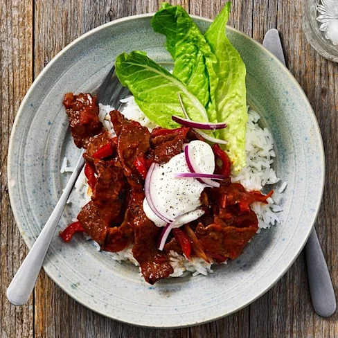 Köttgryta med paprika och lök serverad på ris, garnerad med sallad och en klick gräddfil, på ett träbord.
