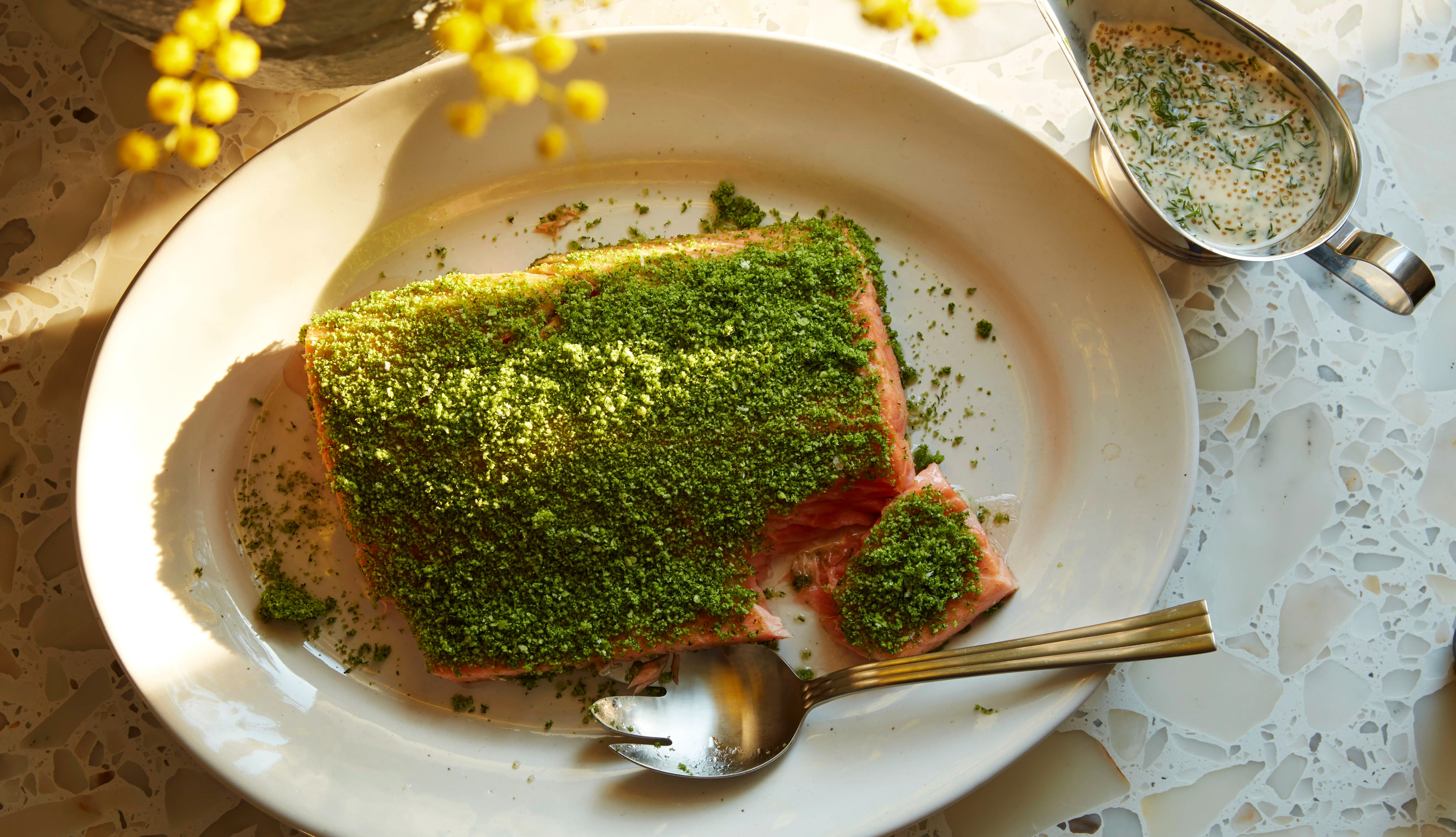 Ugnsbakad lax med grön örtcrust på ett fat, en sked bredvid och en såskanna med dillsås.