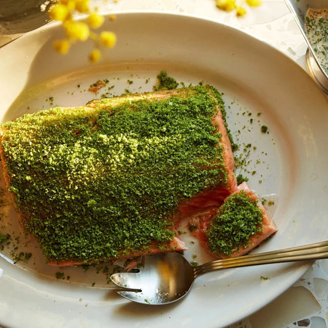 Ugnsbakad lax med grön örtcrust på ett fat, en sked bredvid och en såskanna med dillsås.