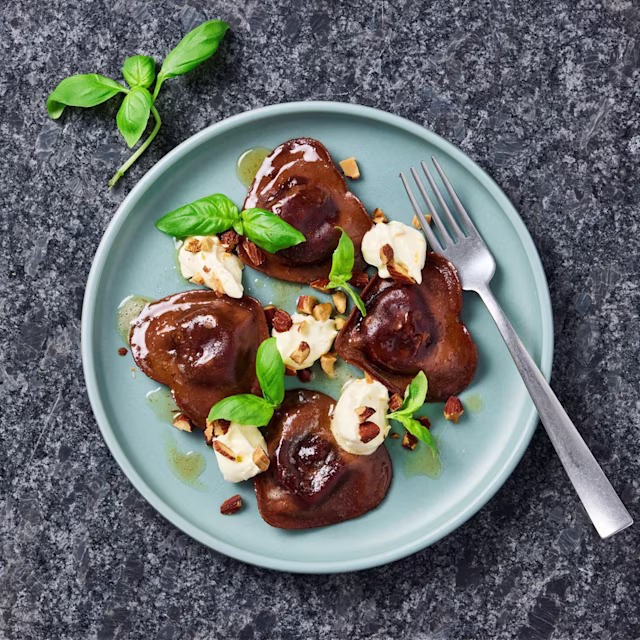 Hjärtformad pasta med chokladfyllning, toppade med gräddfil, nötter och färsk basilika på en blå tallrik.