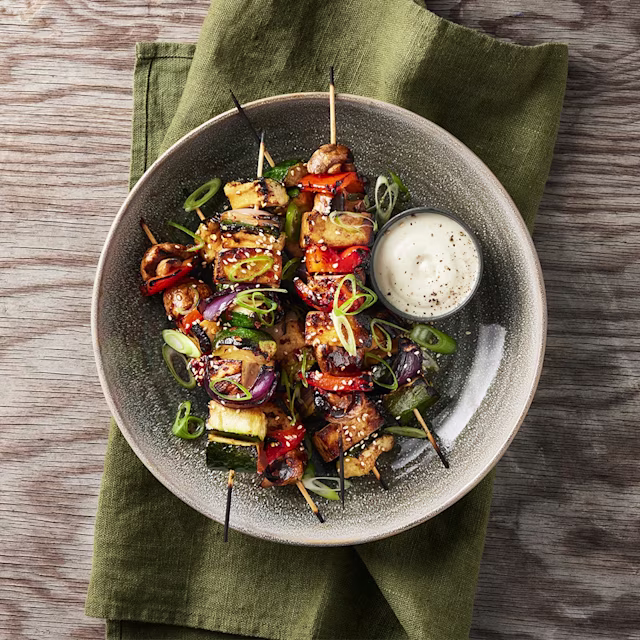 Bild på spett med grönsaker och tofu i en djup tallrik med en liten skål sås.