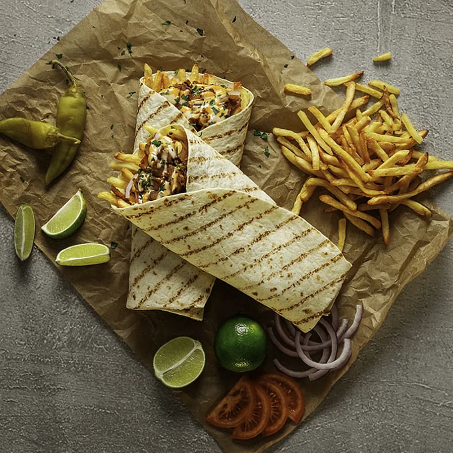 Två wraps med pommes frites och grönsaker, omgivna av limeklyftor, jalapeños, tomatskivor och lök på papper.