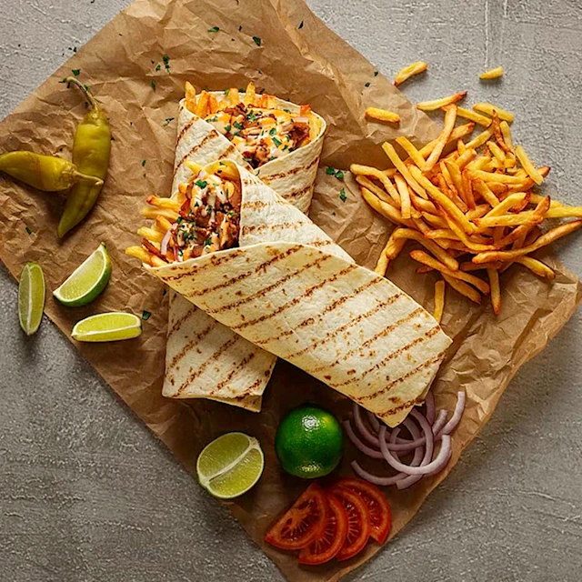 Två wraps med pommes frites och grönsaker, omgivna av limeklyftor, jalapeños, tomatskivor och lök på papper.