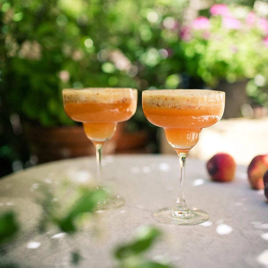 Två cocktailglas med orange drinkar på ett bord utomhus med frukt och gröna växter i bakgrunden.