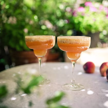 Två cocktailglas med orange drinkar på ett bord utomhus med frukt och gröna växter i bakgrunden.