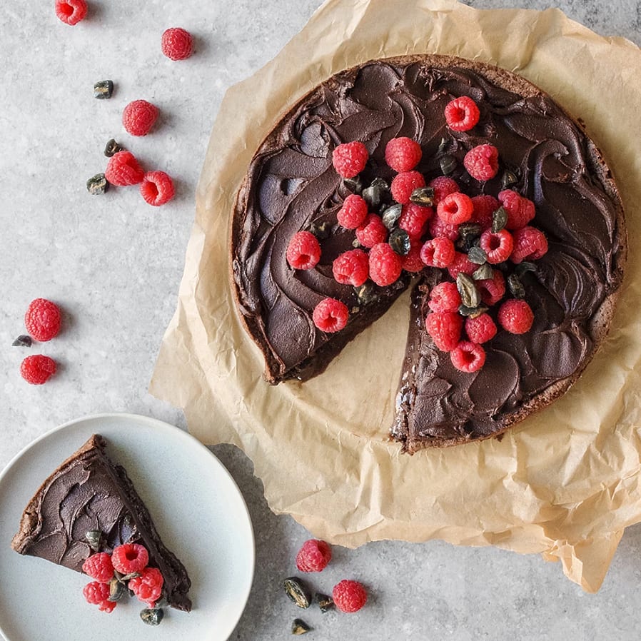En kladdkaka med en tårtbit utskuret. Den utskurna biten kladdkaka ligger på ett fat bredvid. Kakan och tårtbiten är toppad med färska hallon. 