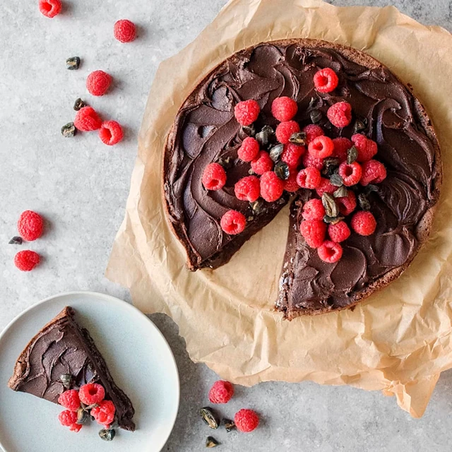 En kladdkaka med en tårtbit utskuret. Den utskurna biten kladdkaka ligger på ett fat bredvid. Kakan och tårtbiten är toppad med färska hallon.