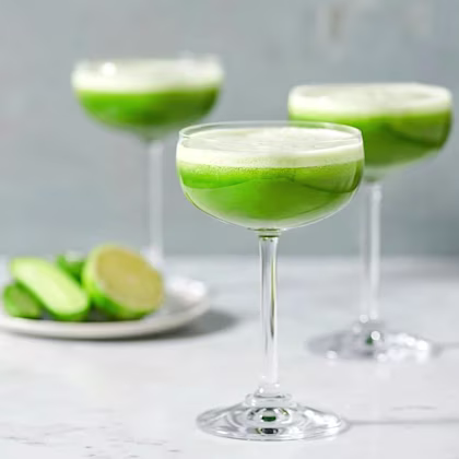 Tre glas med skummig grön cocktail på marmorbord, med limeklyftor på en tallrik i bakgrunden.