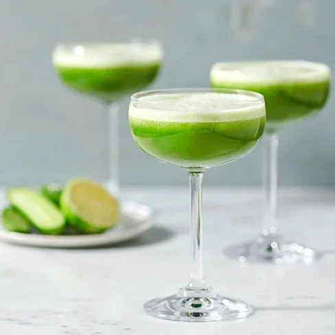 Tre glas med skummig grön cocktail på marmorbord, med limeklyftor på en tallrik i bakgrunden.