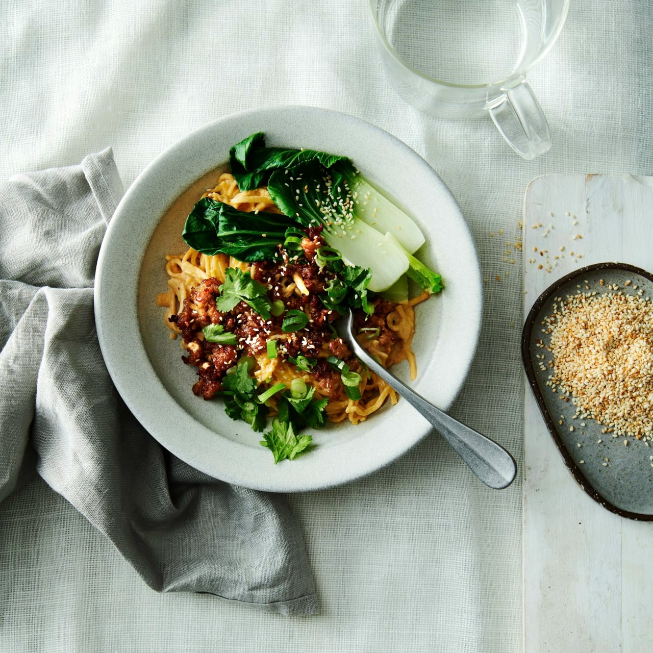 Krämiga nudlar med röd curry och krispig färs, toppade med pak choi, koriander och sesamfrön, serverad i en skål på en duk med en skål sesamfrön och ett glas vatten bredvid.
