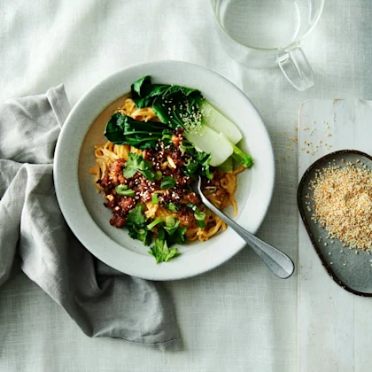 Krämiga nudlar med röd curry och krispig färs, toppade med pak choi, koriander och sesamfrön, serverad i en skål på en duk med en skål sesamfrön och ett glas vatten bredvid.