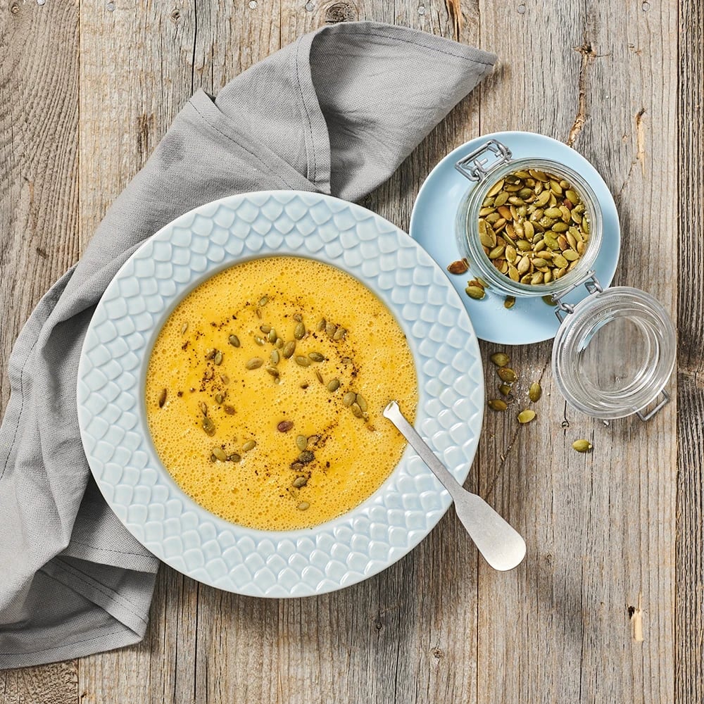 Pumpasoppa toppad med pumpakärnor och kryddor i en djup tallrik, med en sked och en burk pumpakärnor bredvid