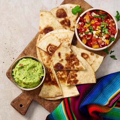 Quesadillas skurna i trekanter på en skärbräda, serverade med guacamole och tomatsalsa i skålar samt en färgglad tygservett.