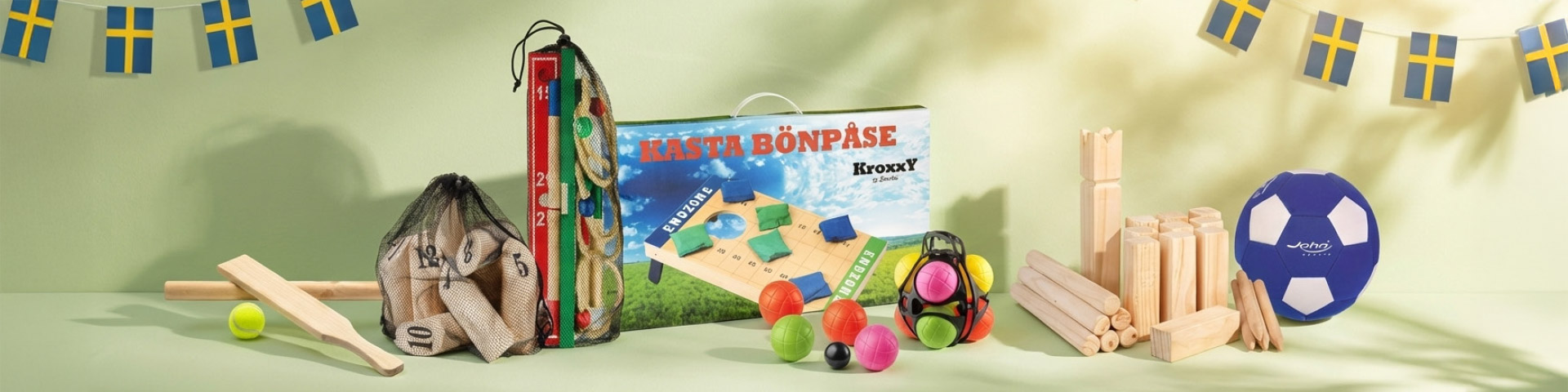 Trädgårdsspel för midsommar med krocket, kubb, kastspel och bollar uppställda på bord med svenska flaggor, med texten “KASTA BÖNPÅSE KroxxY”. Klicka för att handla.