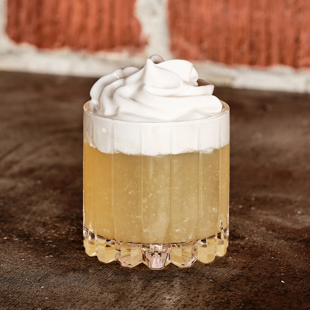 Drink i glas med ljusgul vätska toppad med ett tjockt lager vispad grädde.