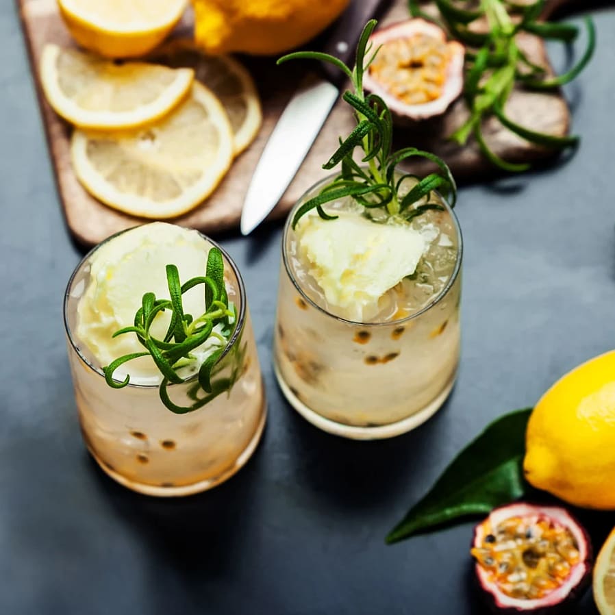Två citrusdrinkar med is, passionsfrukt och rosmarin på ett bord med citroner och skivad citron.