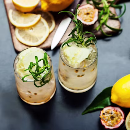 Två citrusdrinkar med is, passionsfrukt och rosmarin på ett bord med citroner och skivad citron.