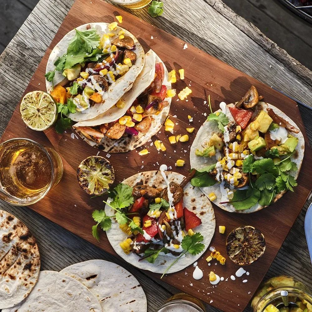 Tacos med grillade grönsaker, majs, avokado, koriander och dressing på tortillabröd, serverade på en skärbräda med grillad lime och dryck bredvid.