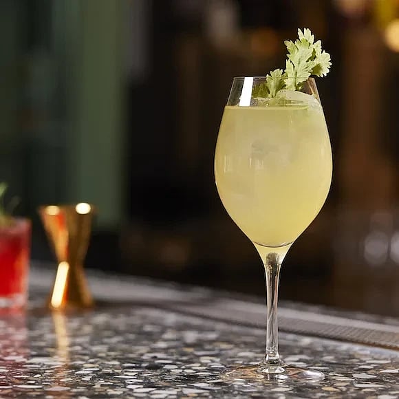 Gul drink i vinglas med is och koriander som garnering stående på marmorbord i bar.