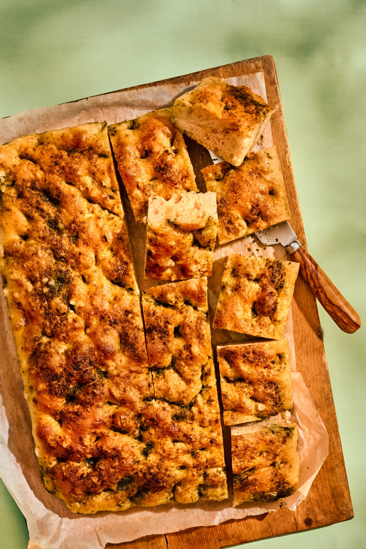 Foccacia med ramslök skuren i rutor på en skärbräda med bakplåtspapper och en kniv.