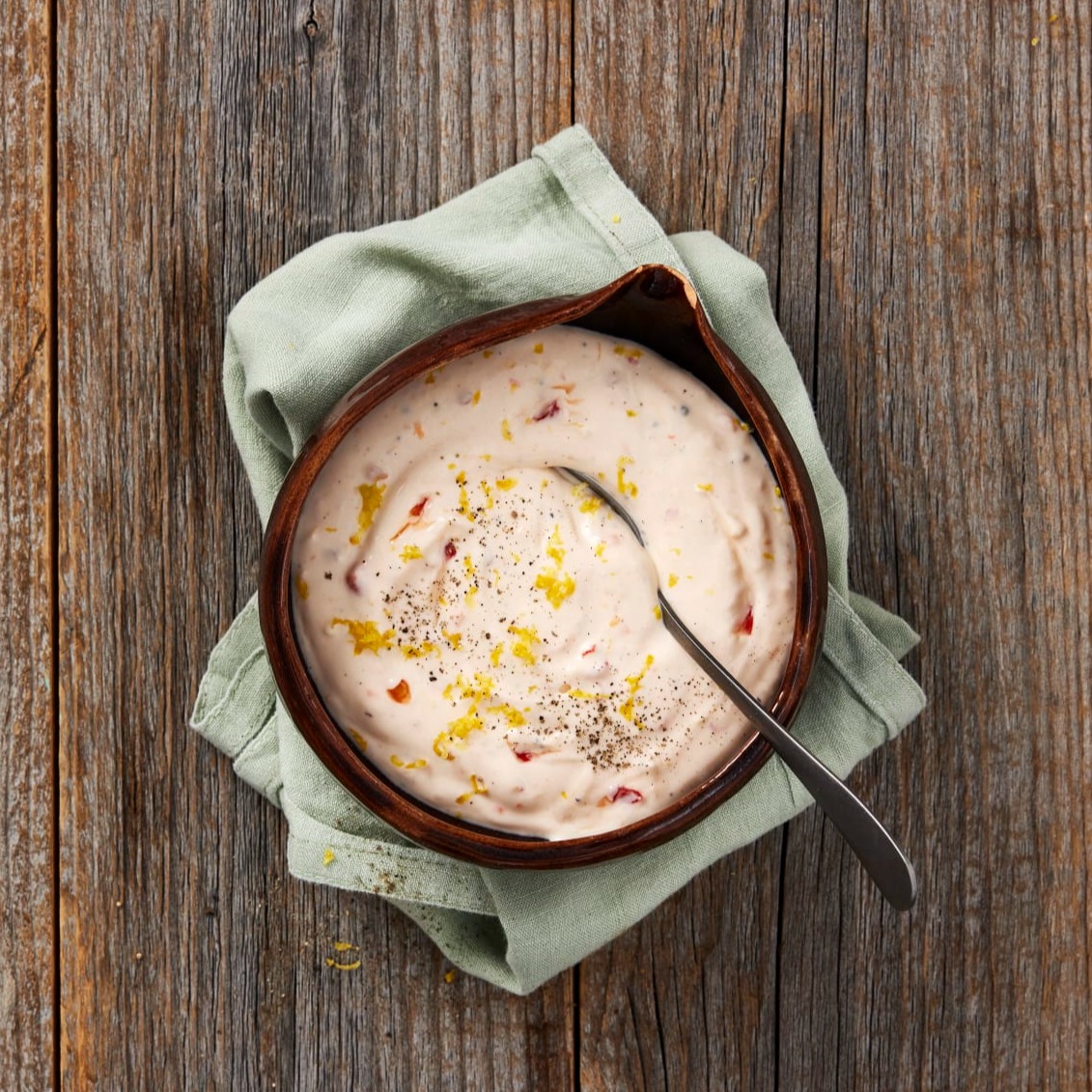 Het yoghurtsås i en skål som står på ett slitet träbord, skålen står på en ljusgrön tygservett. I såsen ligger en sked, såsen är dekorerad med en skvätt olja och svartpeppar. 