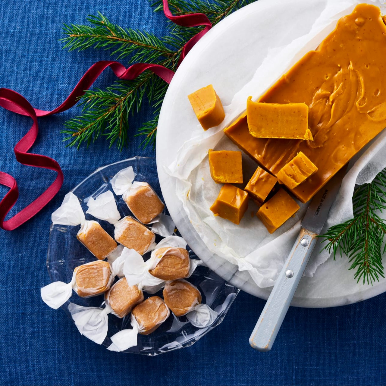 Saffransfudge skuren i bitar på ett fat med kniv och granris, samt inslagen saffransfudge i papper på ett glasfat.