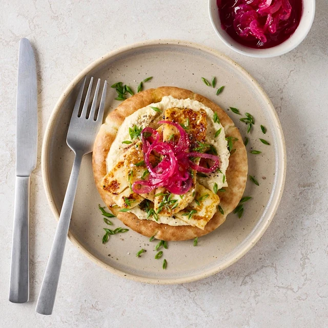 Honungsglacerad tryffelhalloumi med hummus på pitabröd, toppad med picklad rödlök och örter, upplagt på en tallrik med bestick bredvid samt en skål med extra picklad rödlök.