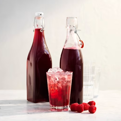 Två flaskor med röd dryck står bredvid ett glas med is och bär, på en ljus yta.