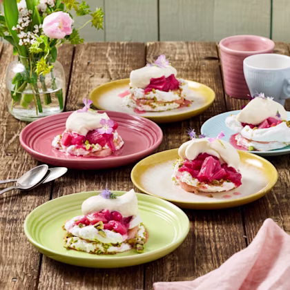 Pavlova med rabarber, grädde och pistagenötter serverad på färgglada assietter, med blommor som dekoration och en vas med blommor i bakgrunden.